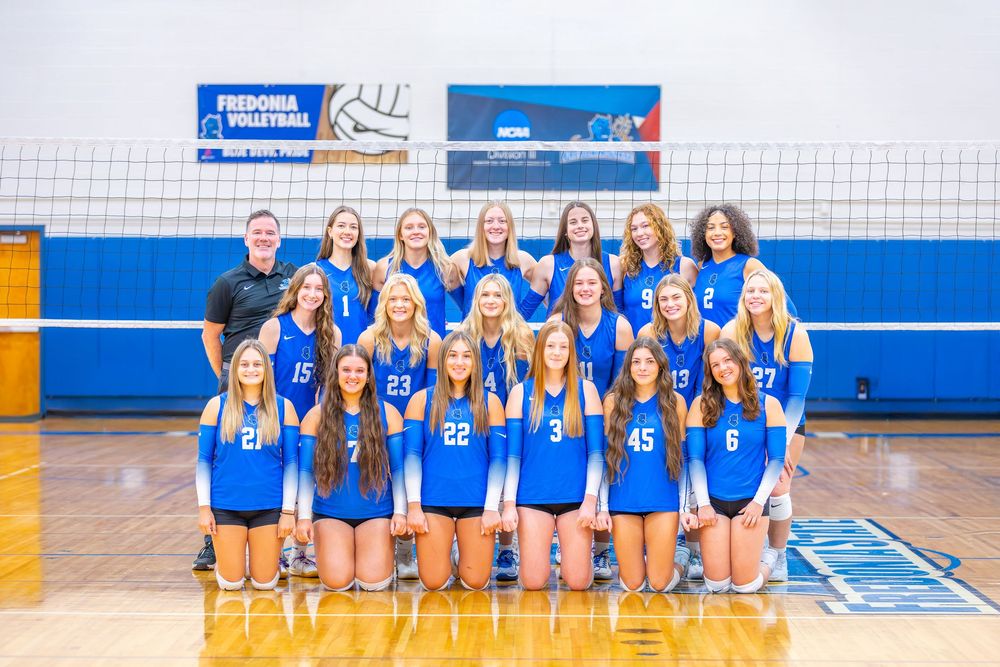 Volleyball team poses for a group photo on the court wearing blue and white uniforms.