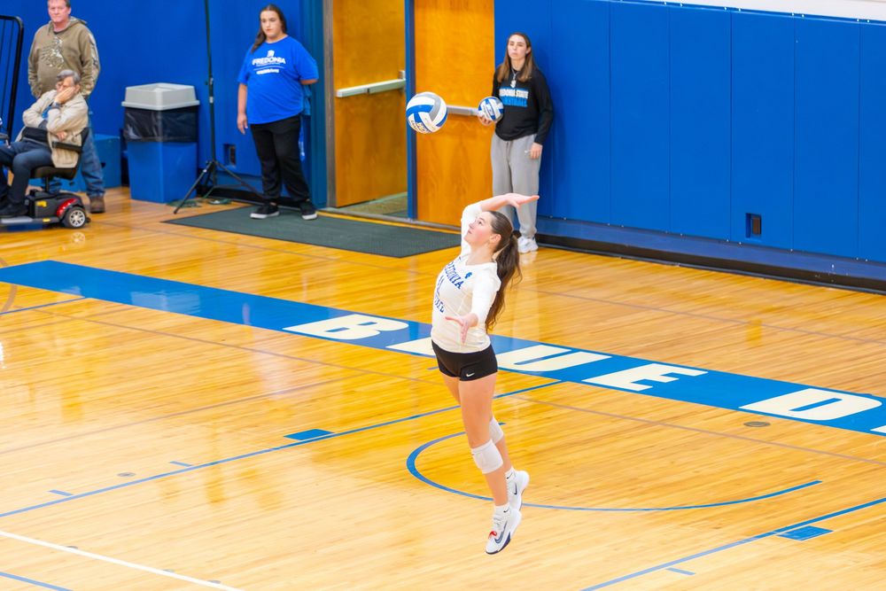 Volleyball player prepares to serve ball during match on blue indoor court.