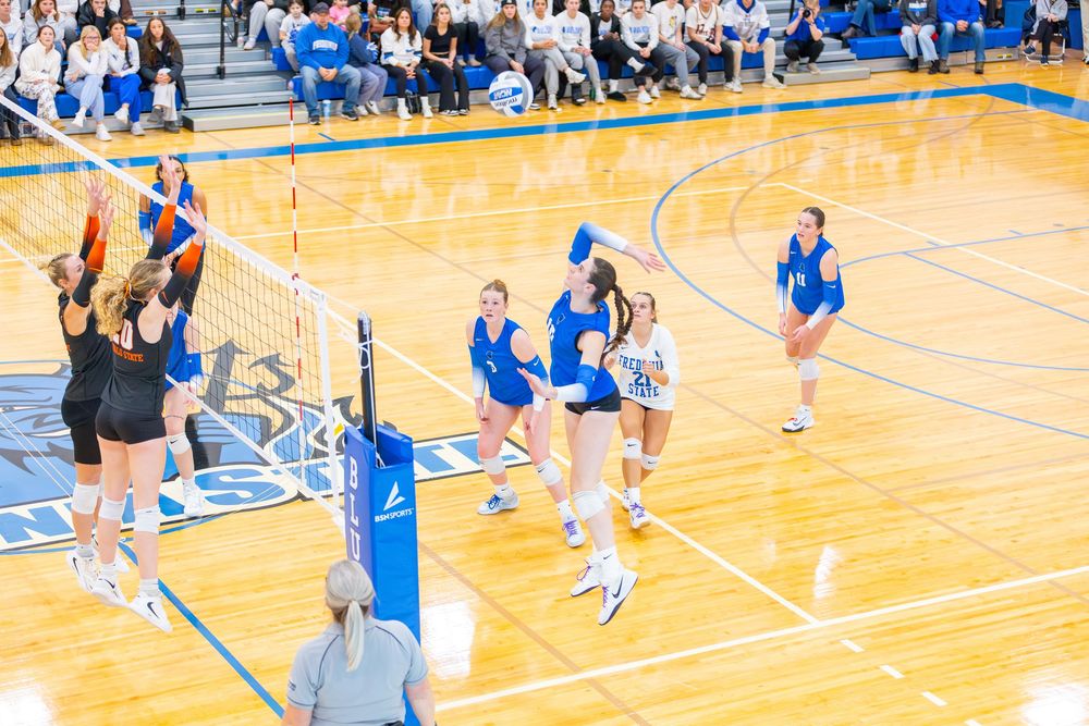 Volleyball players warming up before a match on a bright blue court with spectators watching from the bleachers.