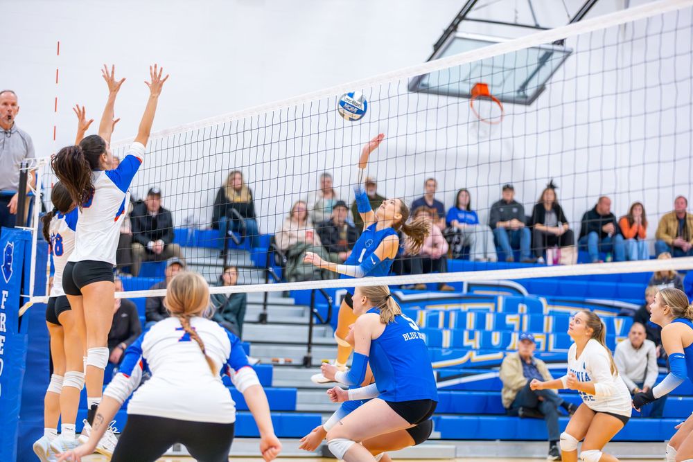 Volleyball players leap at the net during an intense match in a blue and white gymnasium.