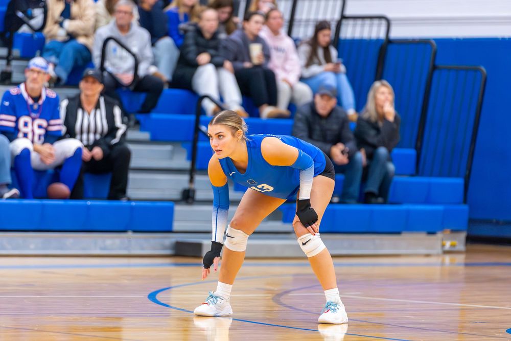 Volleyball player in blue and white uniform crouches in defensive position on indoor court during match.