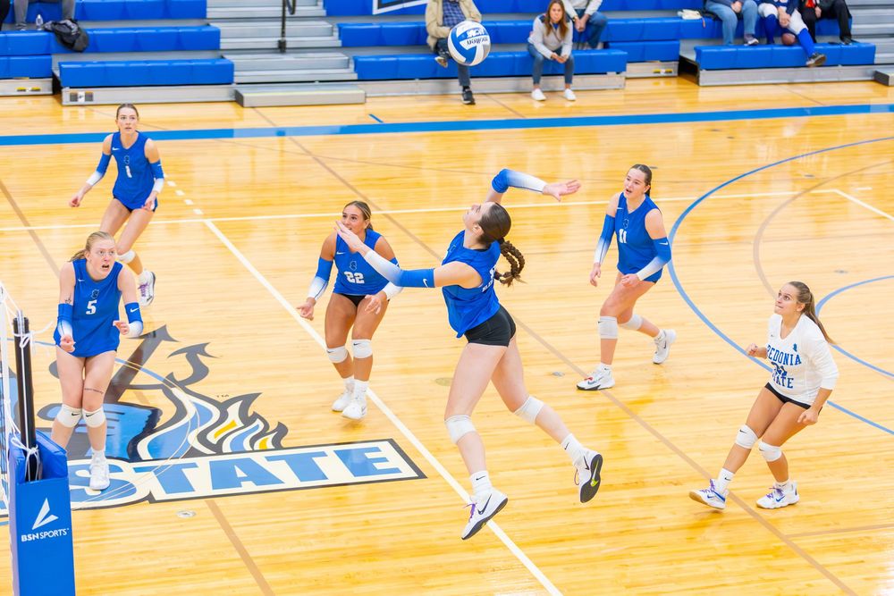 Multiple volleyball players in blue uniforms practice defensive drills on a bright indoor court.