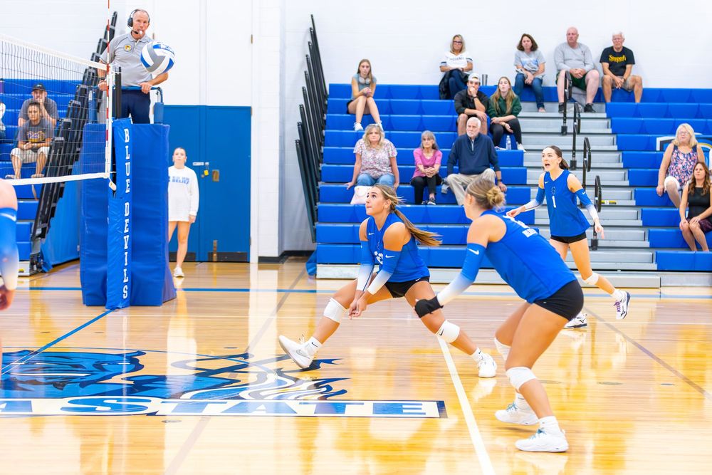 High school volleyball players in blue and white uniforms compete during an intense match on a gymnasium court.