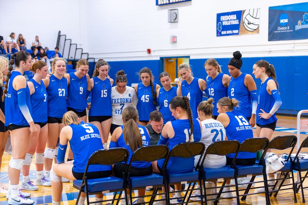 High school volleyball team sits on chairs along sideline during game in blue and white gymnasium.