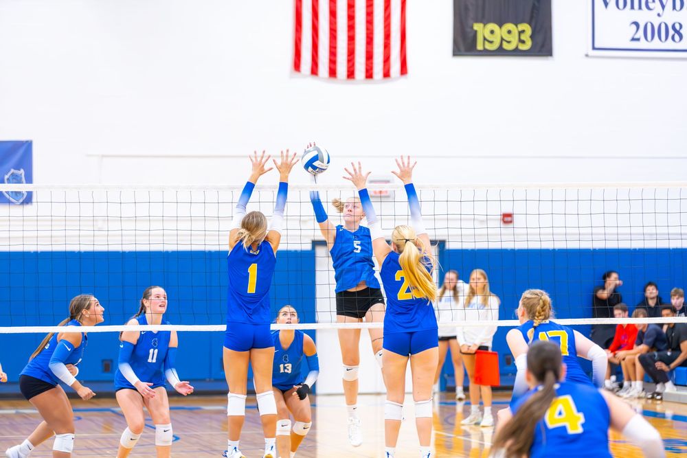 Indoor volleyball players in blue uniforms jump to block a spike during an intense match.