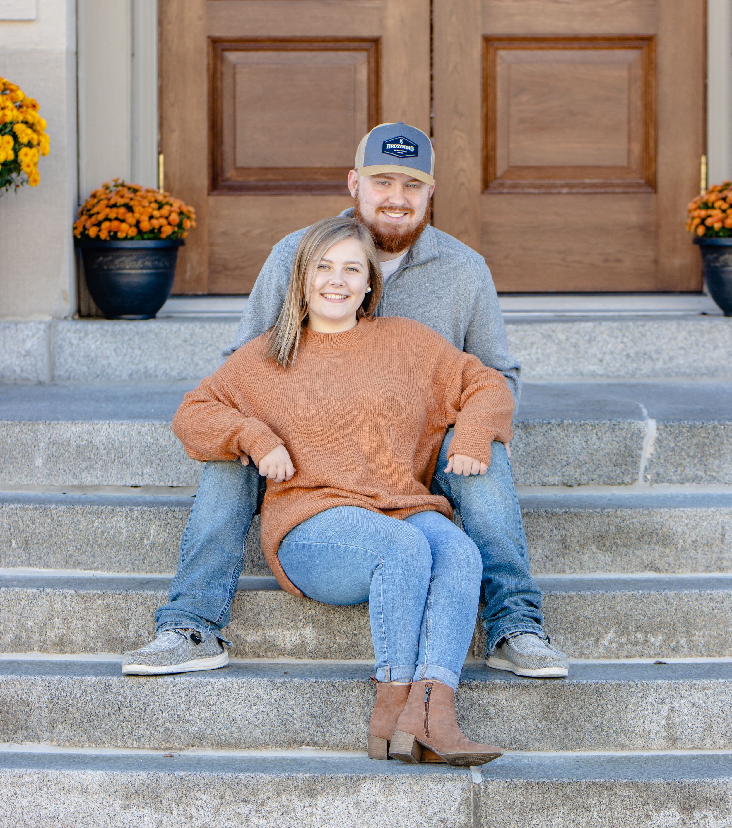 A couple sits casually on concrete steps with fall decorations and orange flowers nearby.