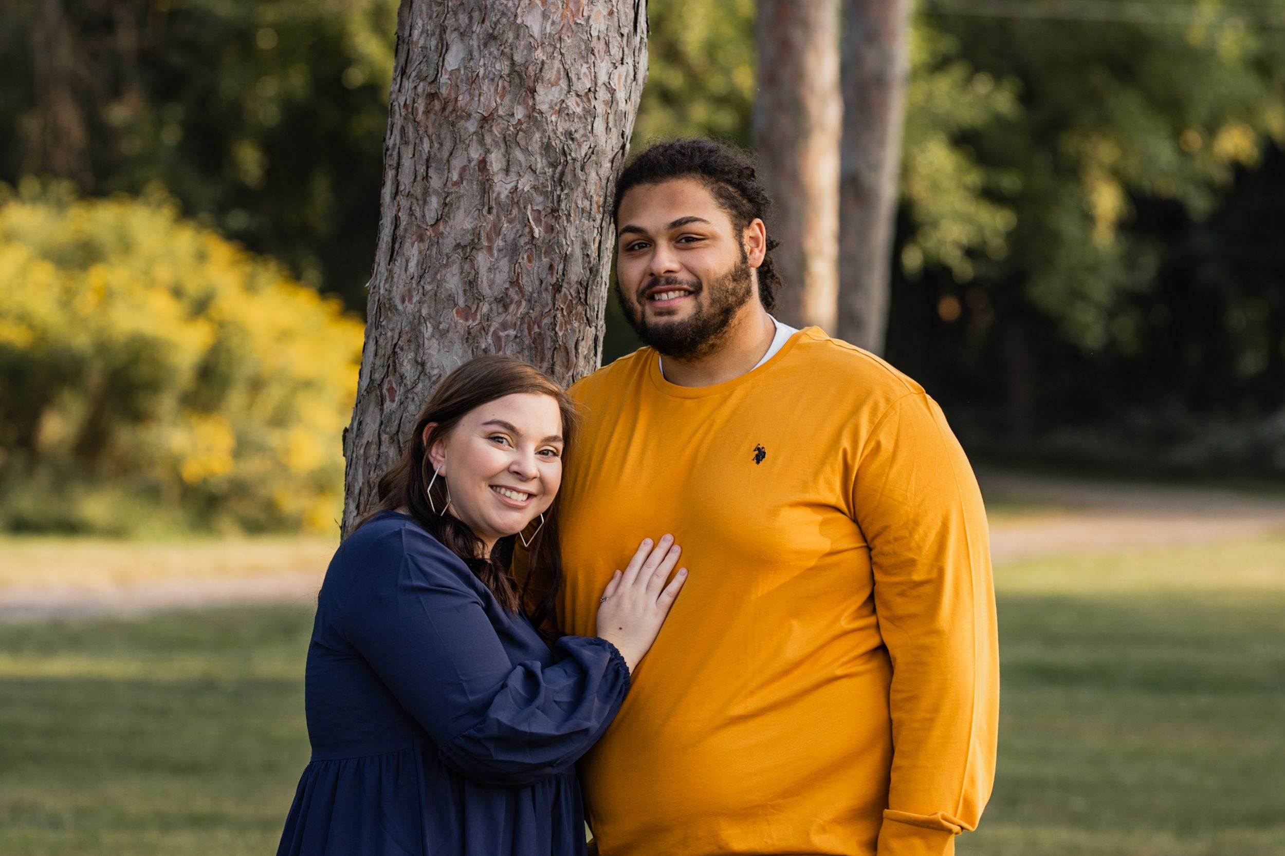 A couple embraces near a tree in a park setting with autumn colors in the background.