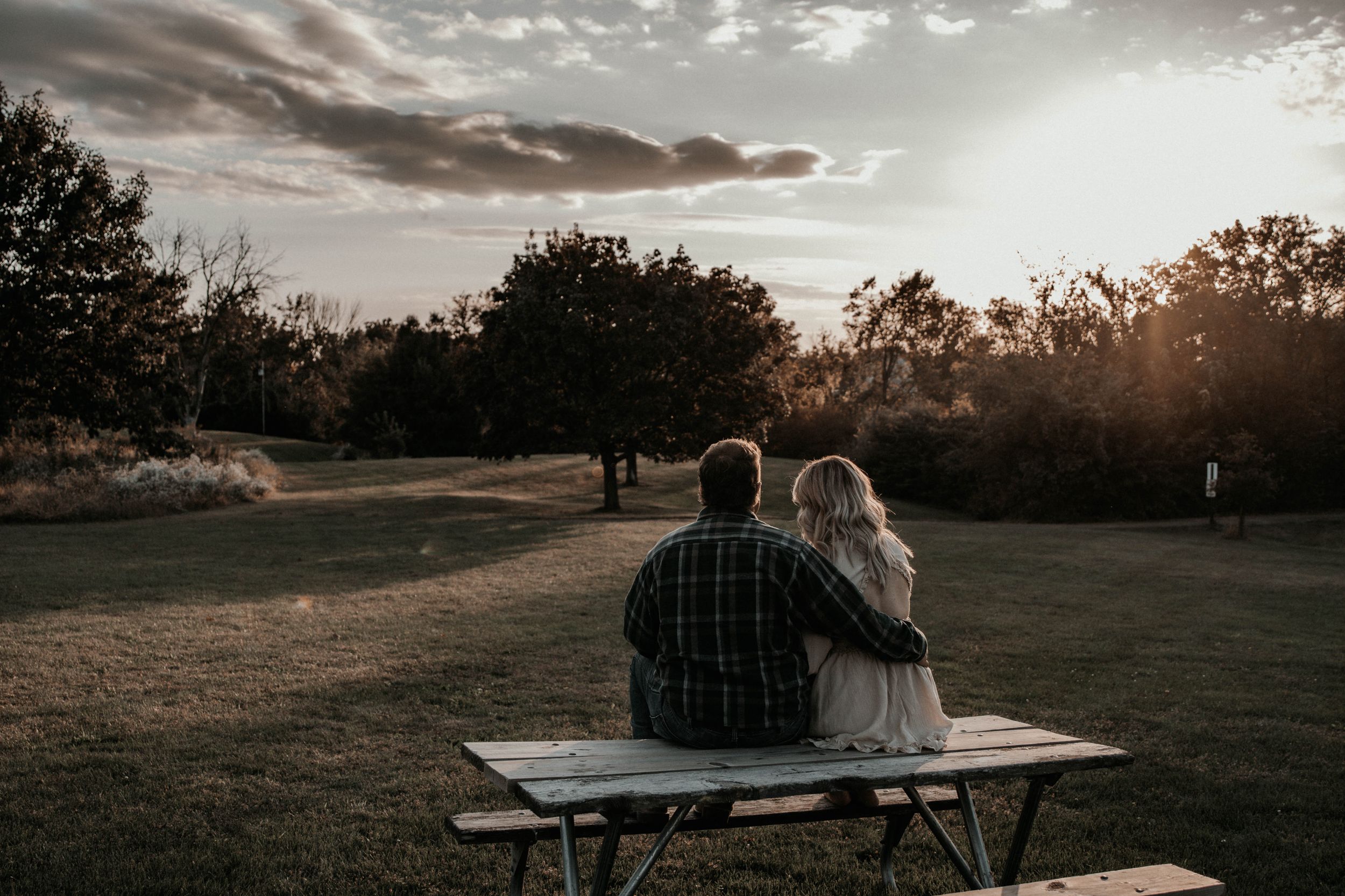 Two people sitting on bench watching dramatic sunset over open field.
