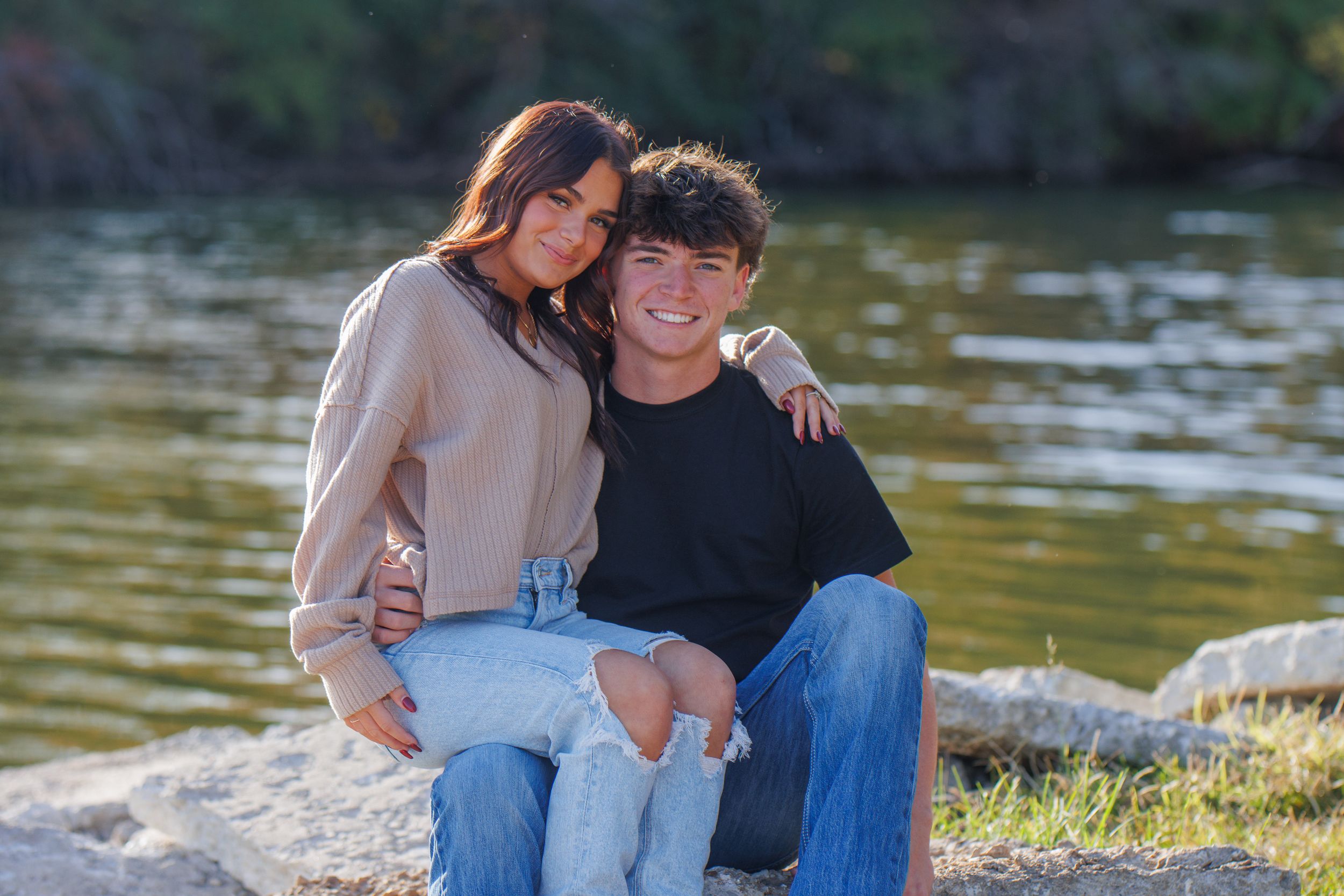 A couple sits together on rocks near a lake during golden hour.