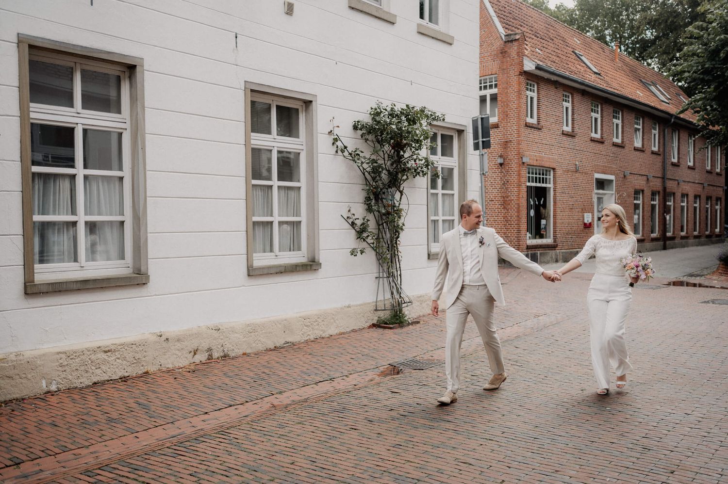 A couple dressed in white holding hands while walking down a brick-paved street past historic European buildings.