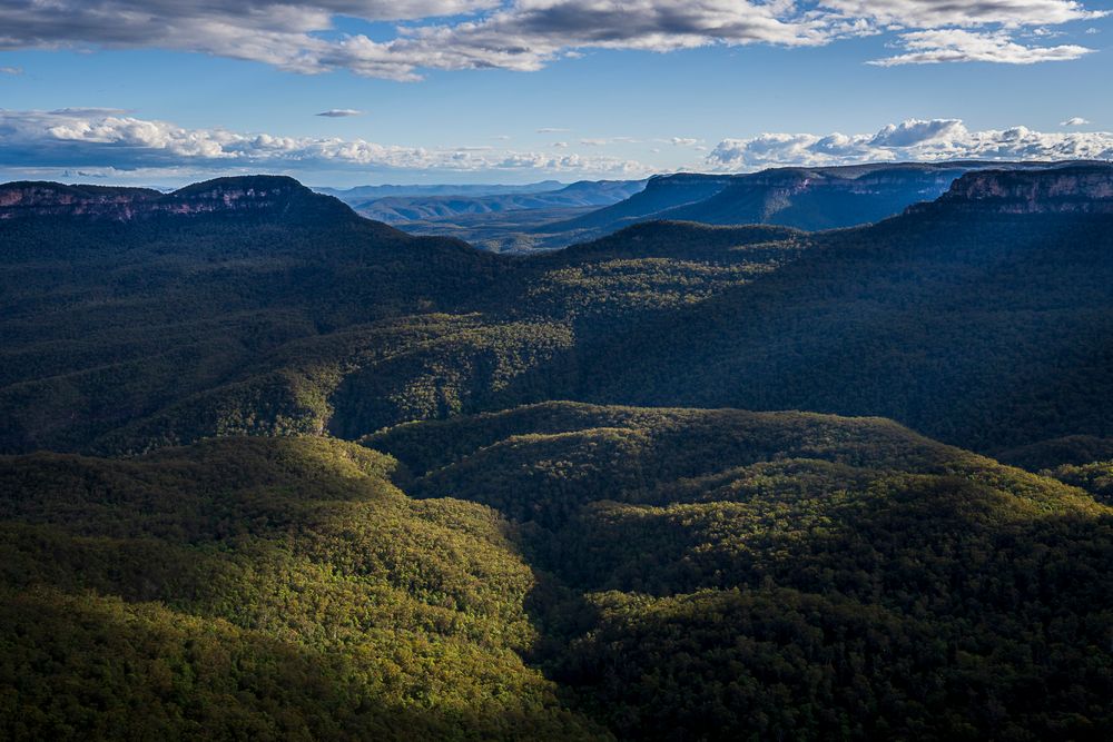 Aerial view of lush green valleys and dramatic sandstone cliffs in the Blue Mountains, Australia, under a partly cloudy sky.
