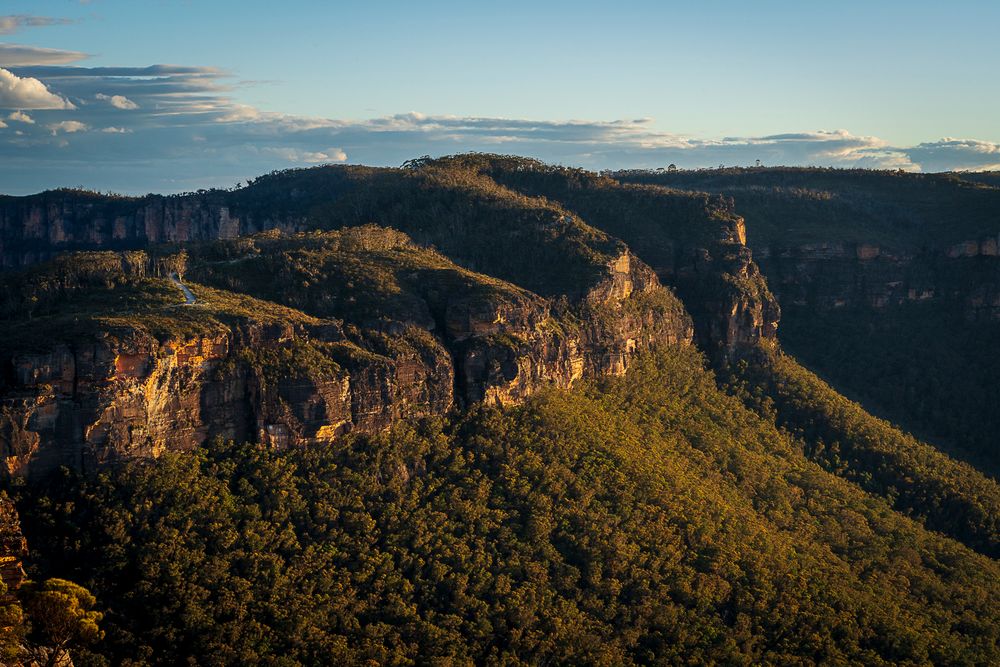 Aerial view of lush green forested cliffs and rocky escarpments in the Blue Mountains, Australia, bathed in golden sunset light.