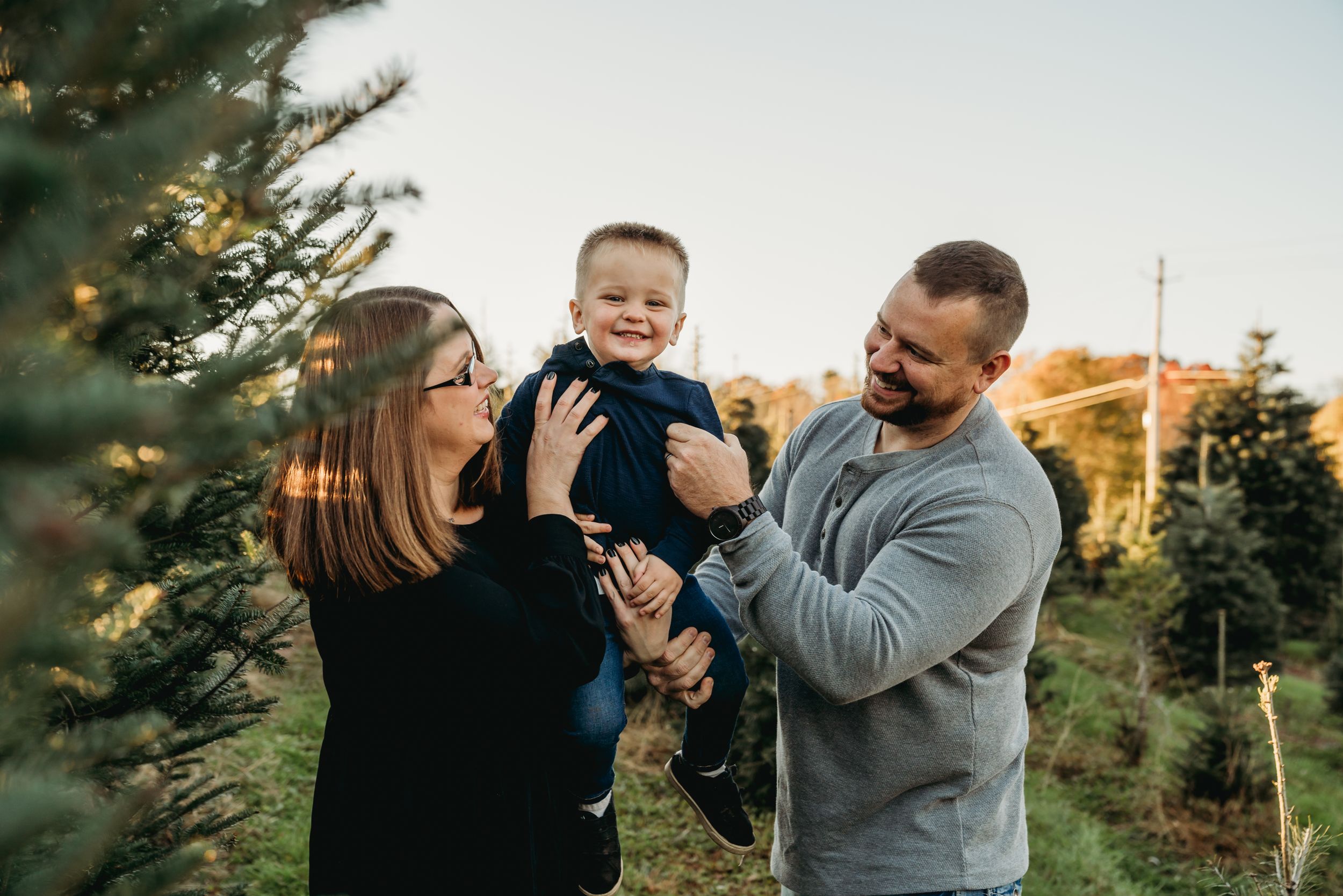A loving family moment captured outdoors among evergreen trees at sunset.