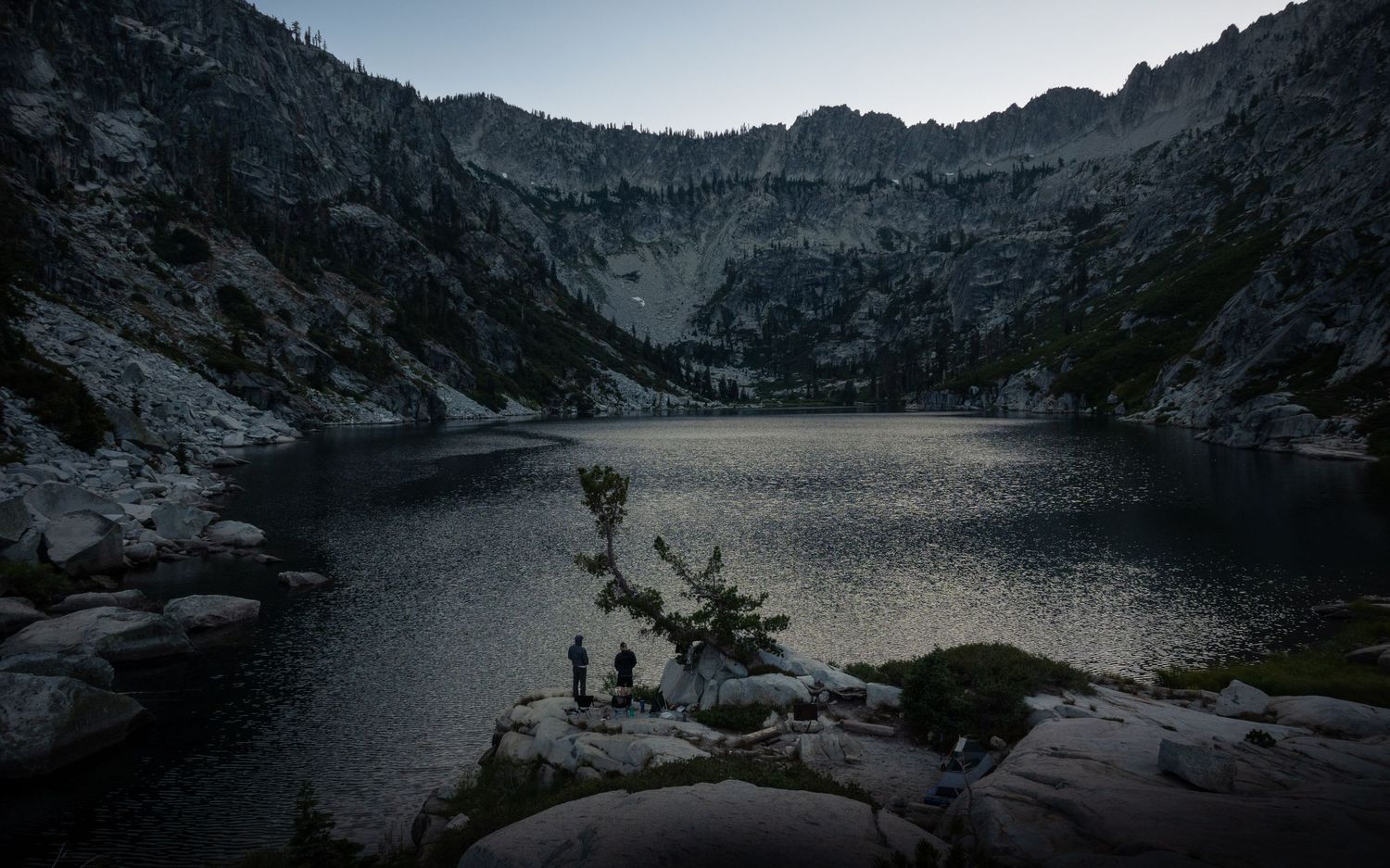 Alpine lake at twilight surrounded by steep rocky walls and scattered boulders.