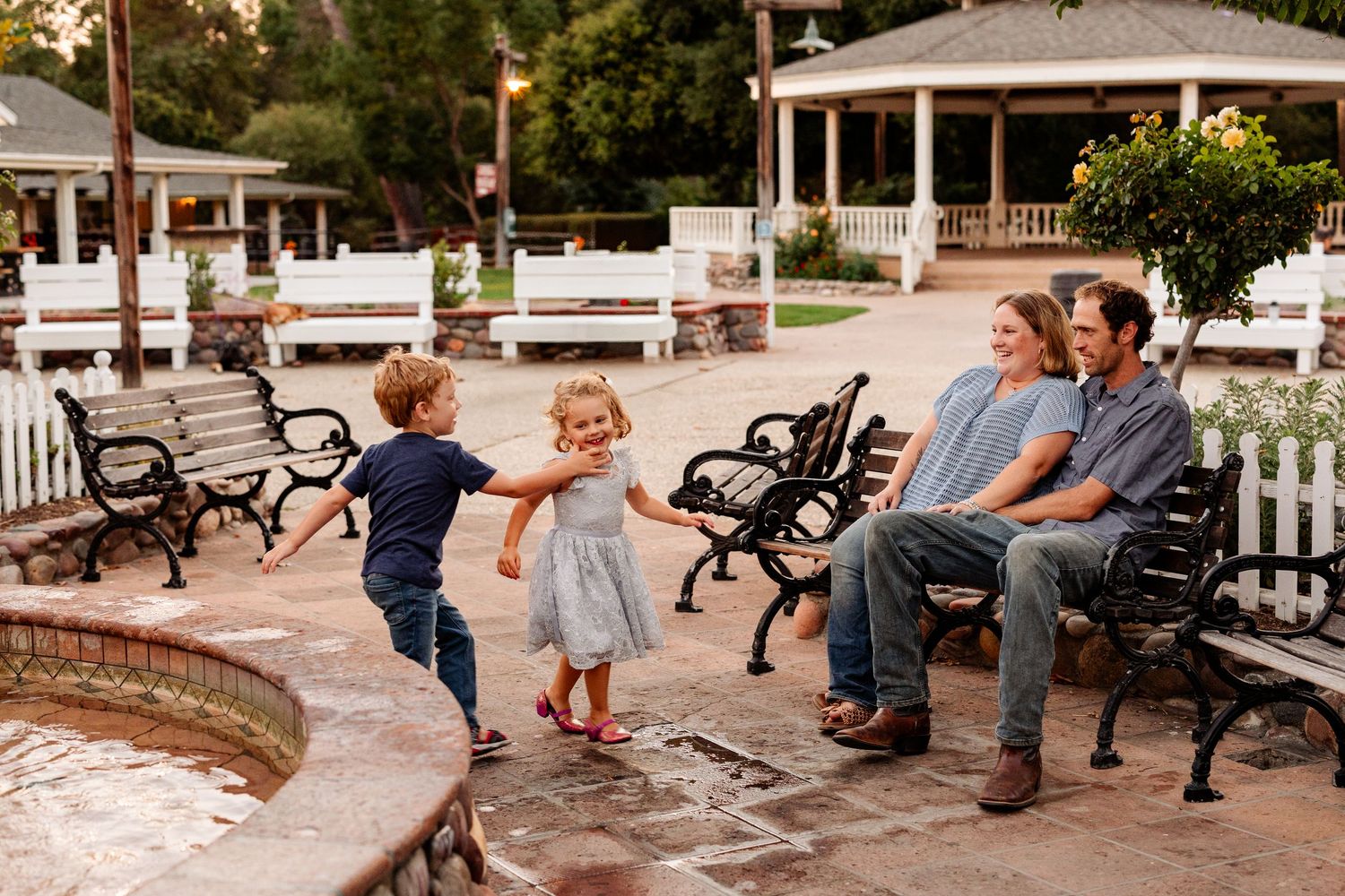 A candid family photo at sunset in Old Poway Park, San Diego. Two young children joyfully play near a fountain while their parents smile.