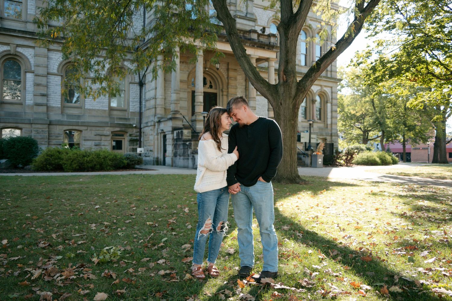 Couple holds hands and smiles at each other on grass in front of historic building in fall.