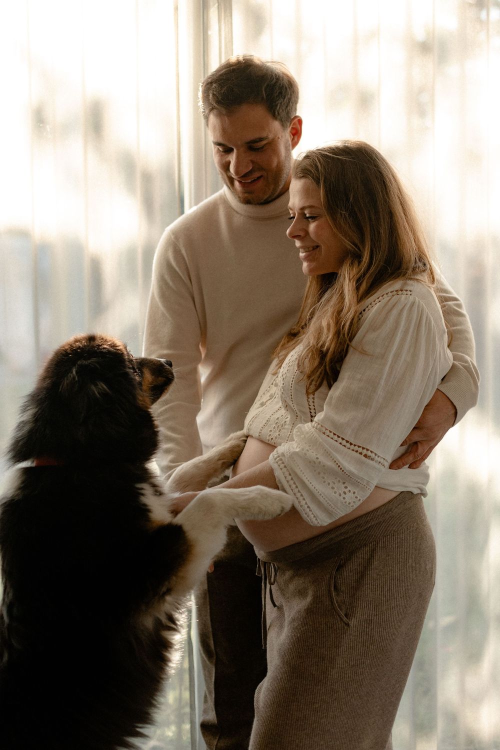 A couple shares tender moments while their black dog interacts with them by a window.