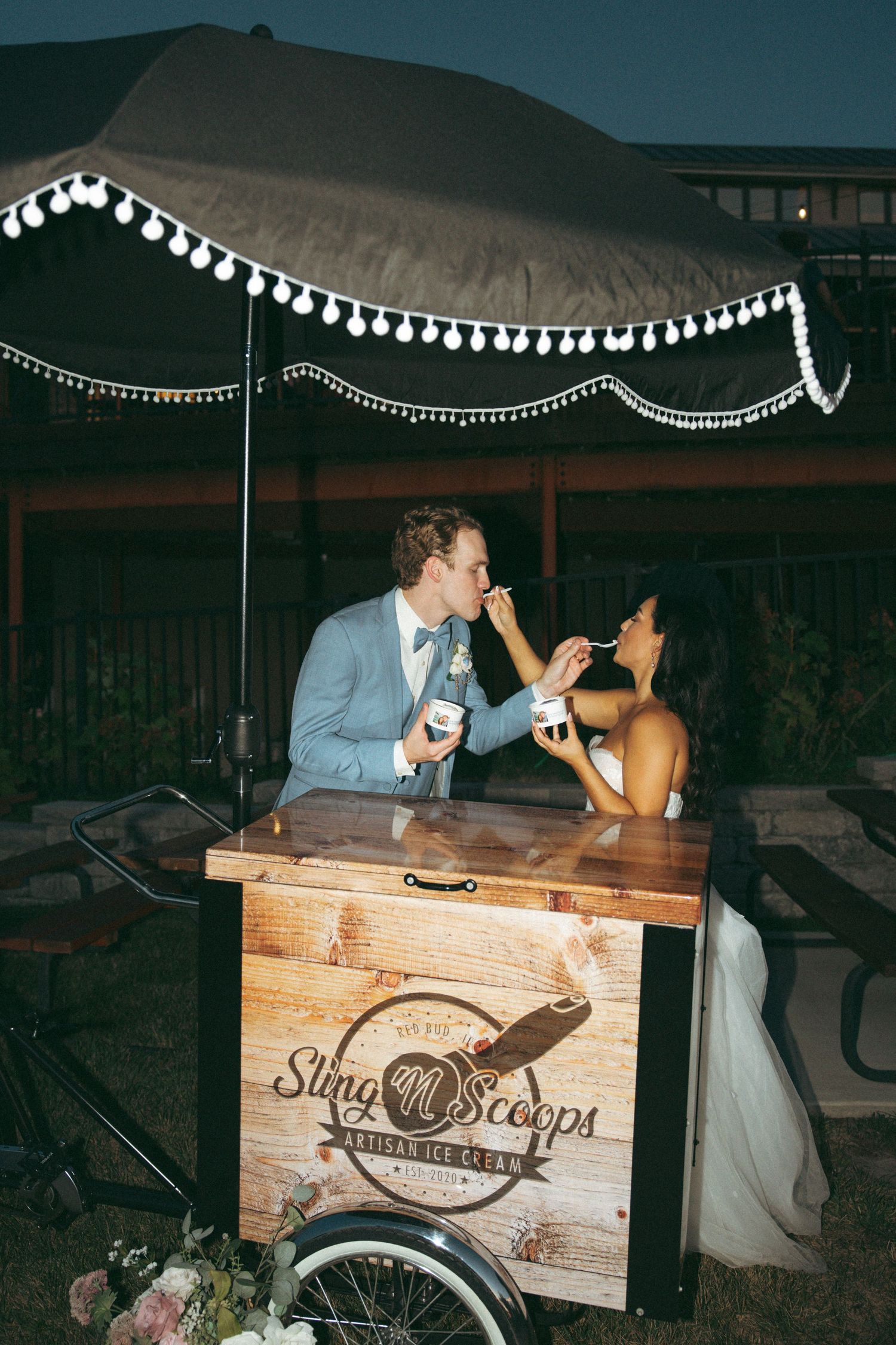 Couple shares ice cream at rustic wooden dessert cart under string lights.