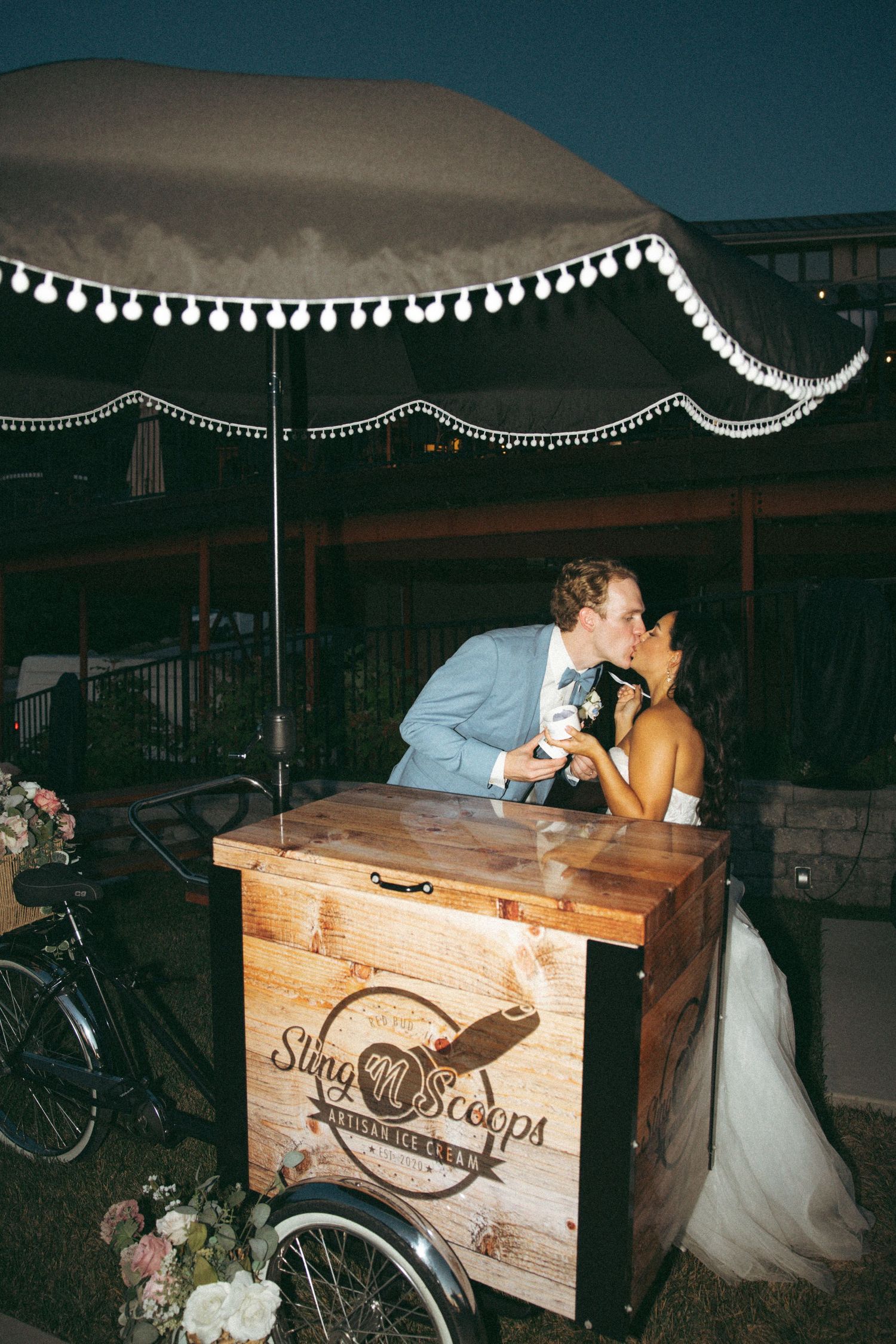 Evening wedding moment at a vintage dessert cart under twinkling pom-pom lights.