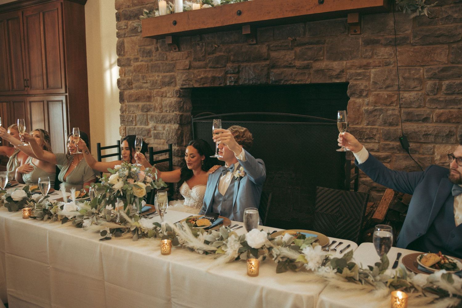 Reception dinner table decorated with floral centerpieces against stone fireplace.
