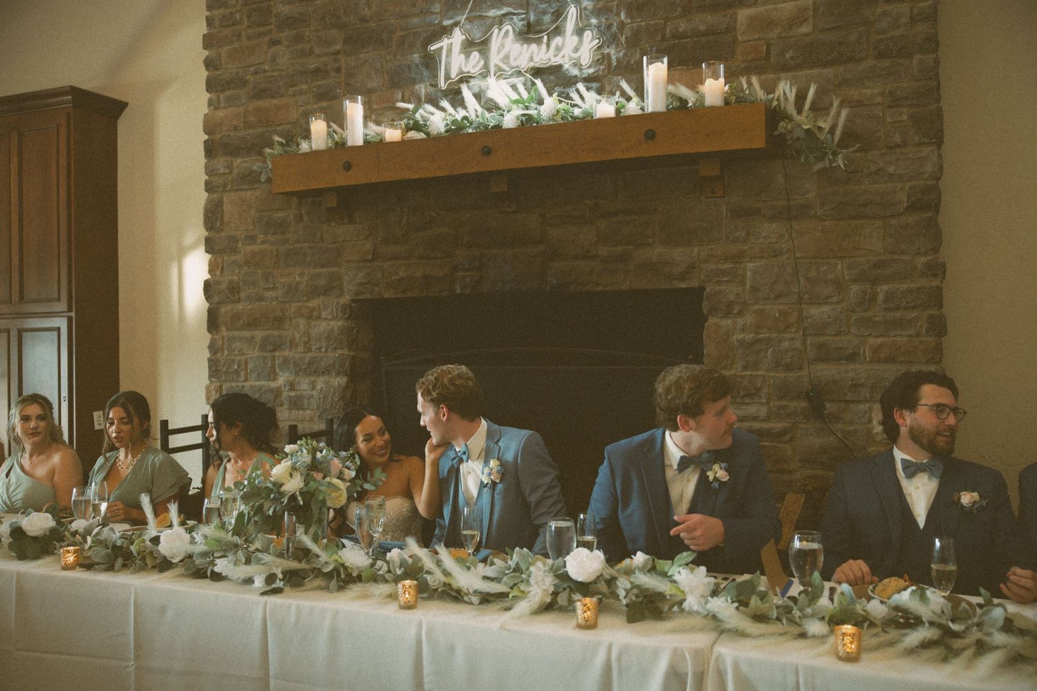 Wedding party seated at a long head table with stone fireplace backdrop and floral decorations.
