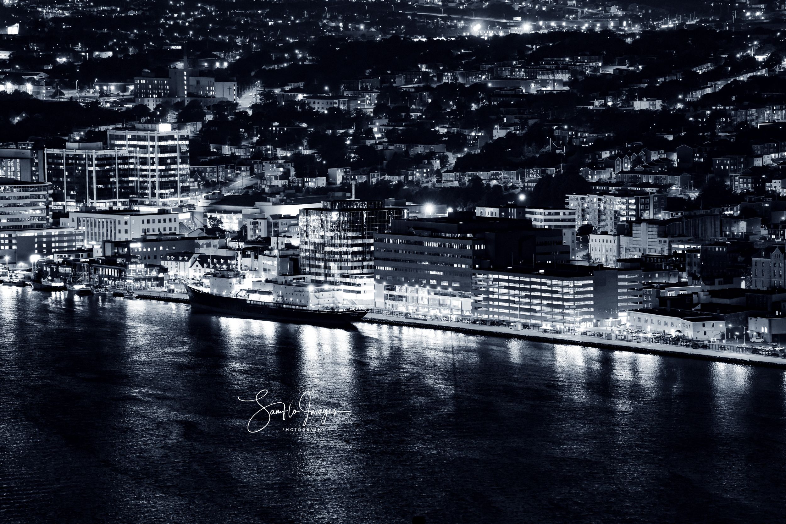 Night view of illuminated waterfront buildings and boats along a harbor in black and white.