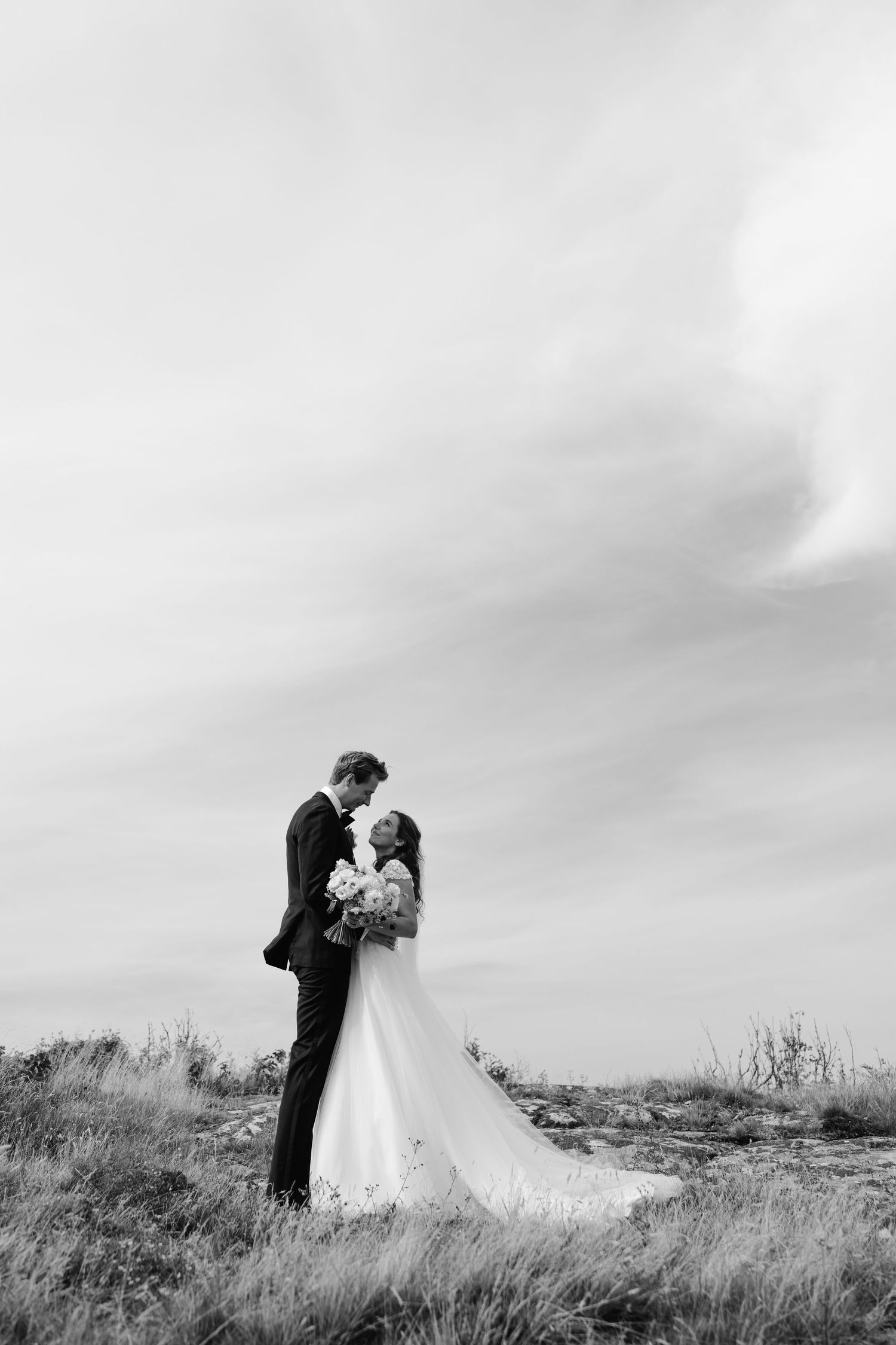 A romantic black and white photo of newlyweds embracing on a hilltop against a dramatic cloudy sky.