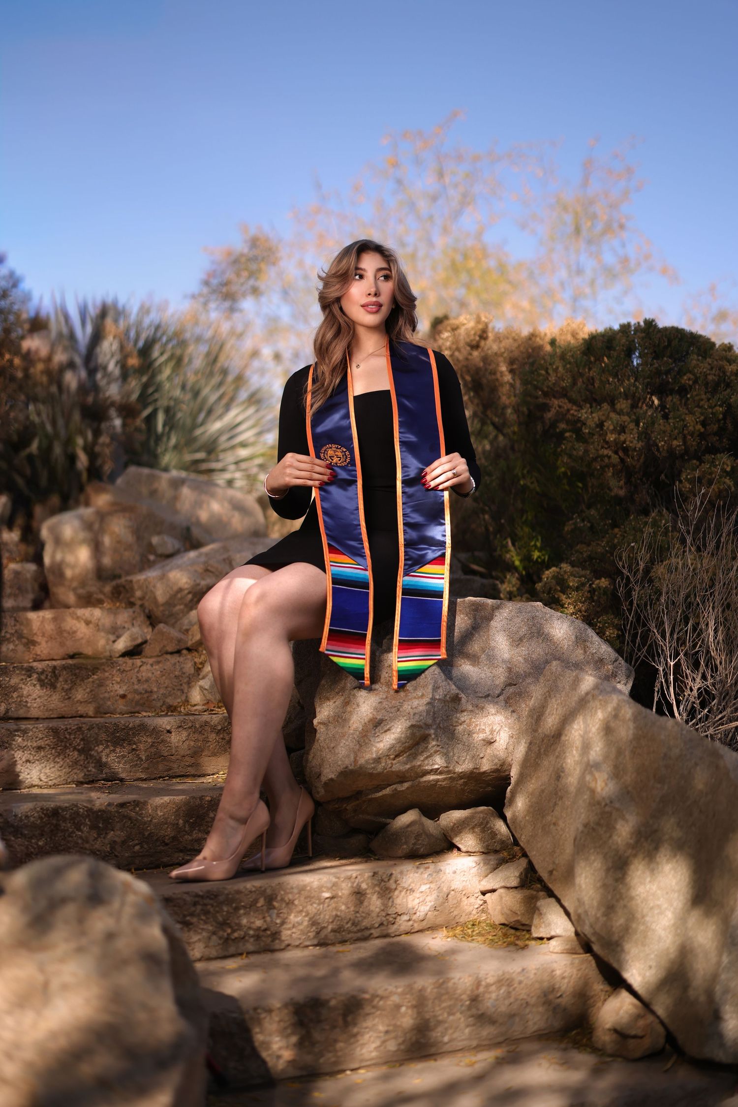 Graduate in black dress with colorful graduation stole sits on natural rock formation during golden hour outdoor photoshoot.