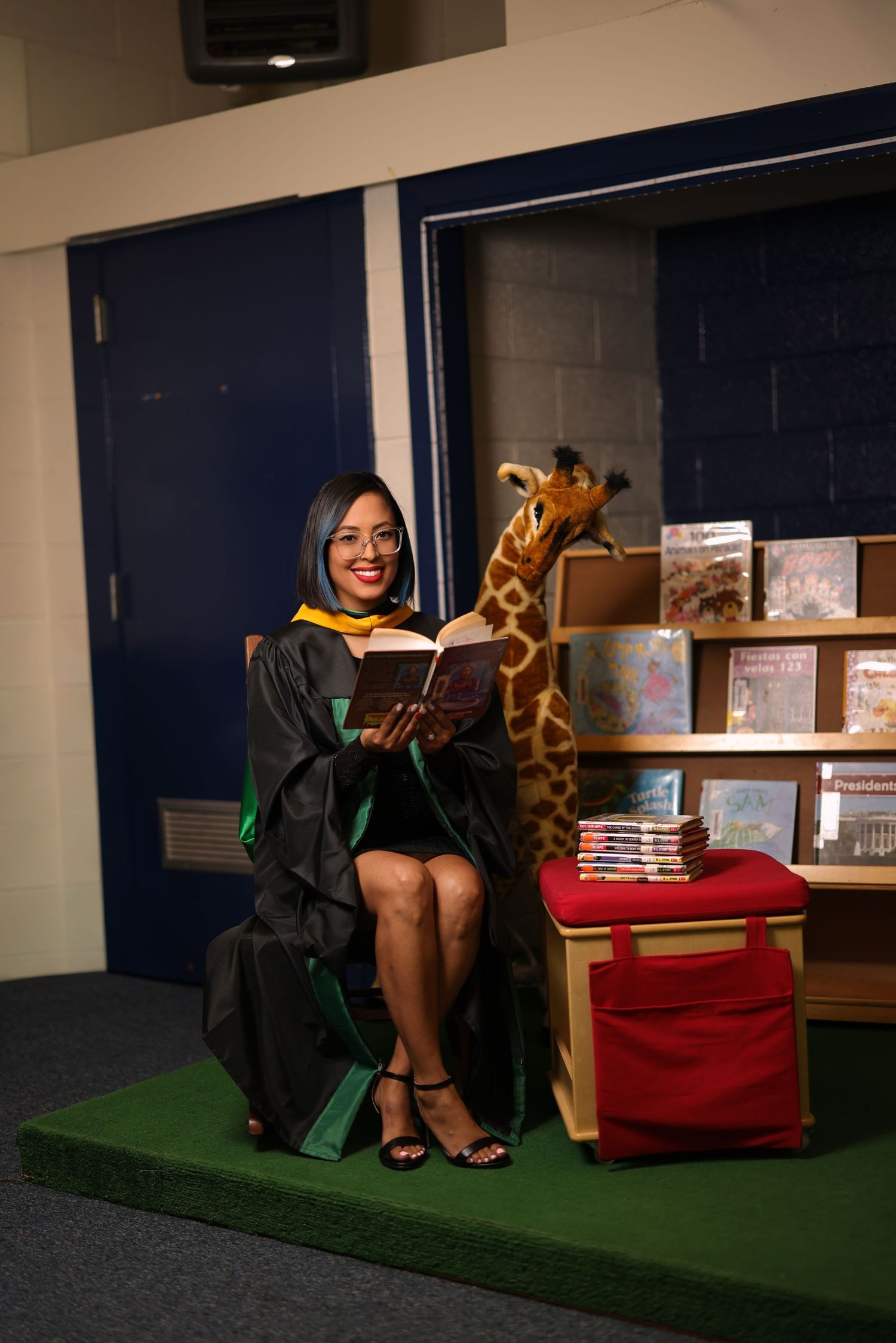A graduate in academic regalia poses with a stuffed giraffe in a classroom setting.