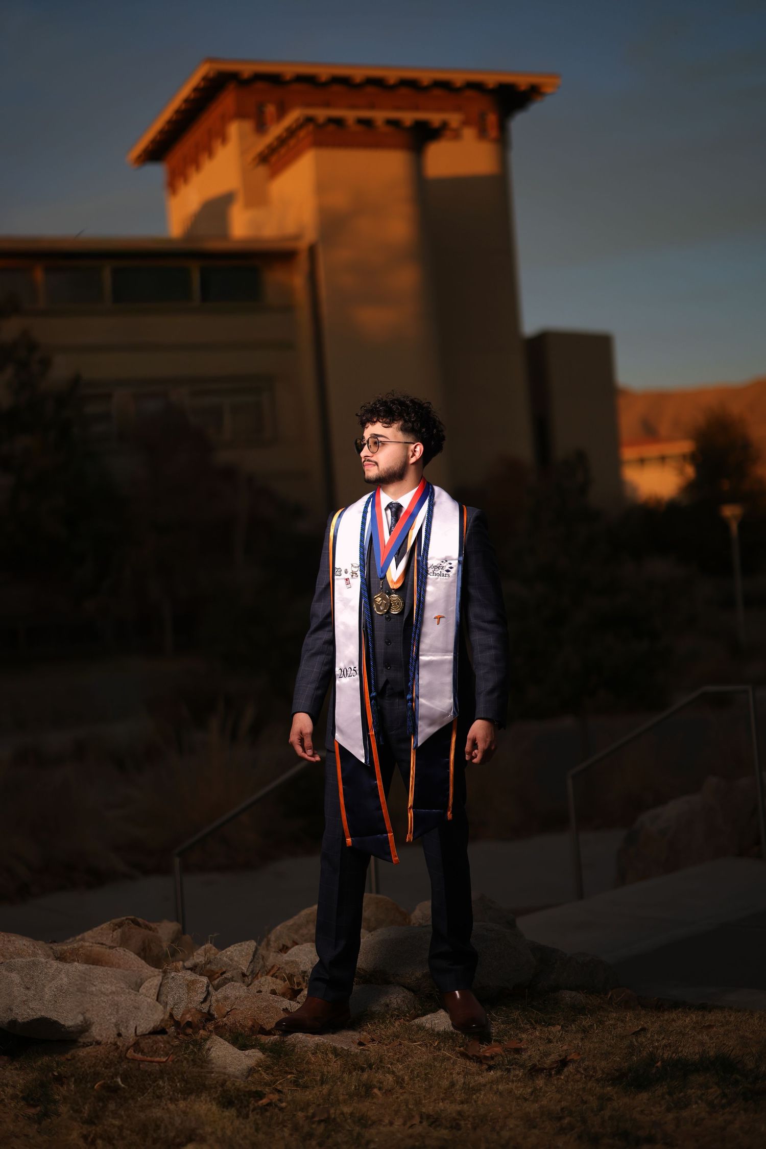 Graduate in cap and gown poses at sunset on rocky terrain wearing graduation stoles and sashes.