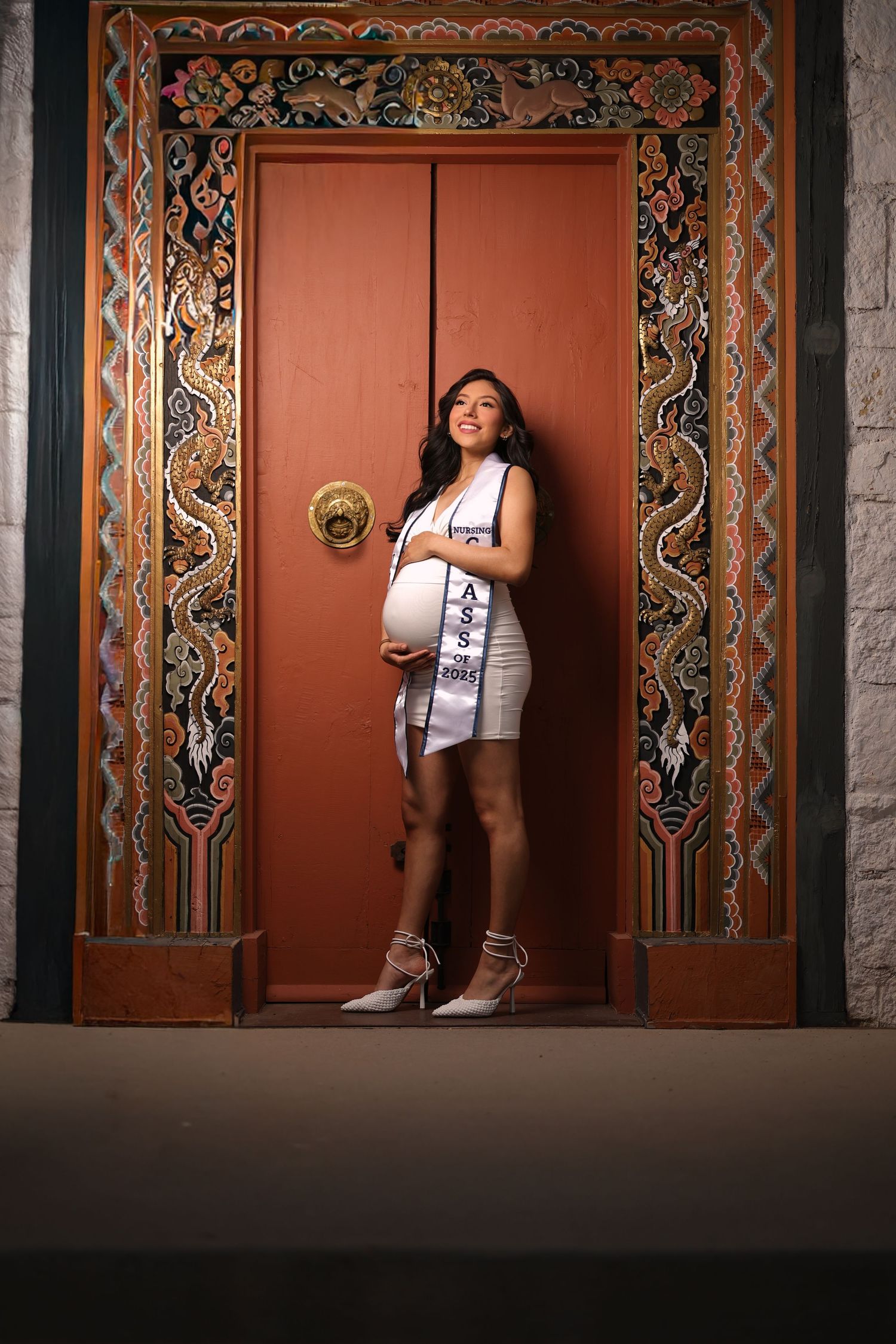 A person poses in a white dress against an ornate doorway with colorful traditional Balinese carved wooden frame details.