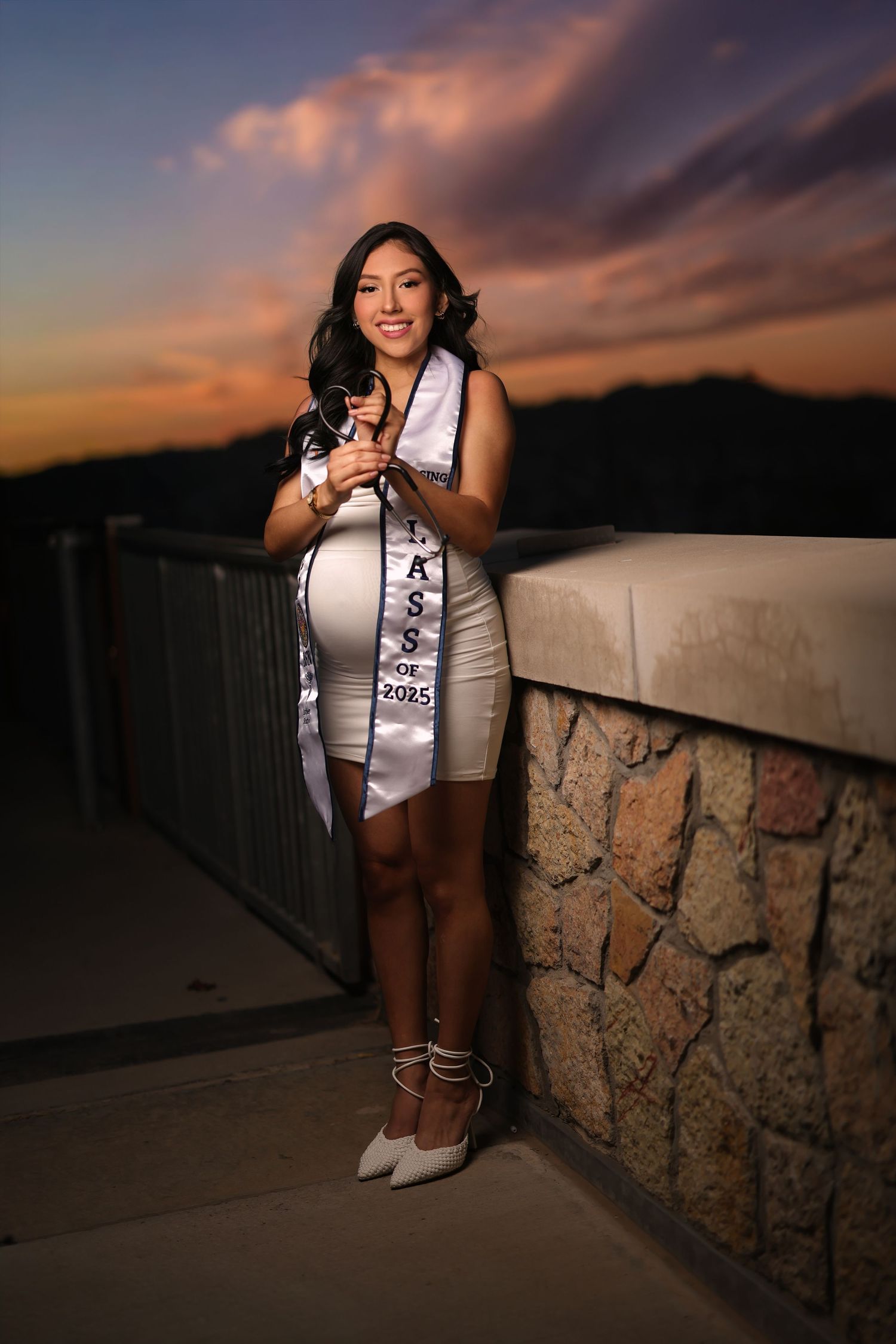 A graduate poses against a stone wall during sunset wearing a white top and graduation sash.