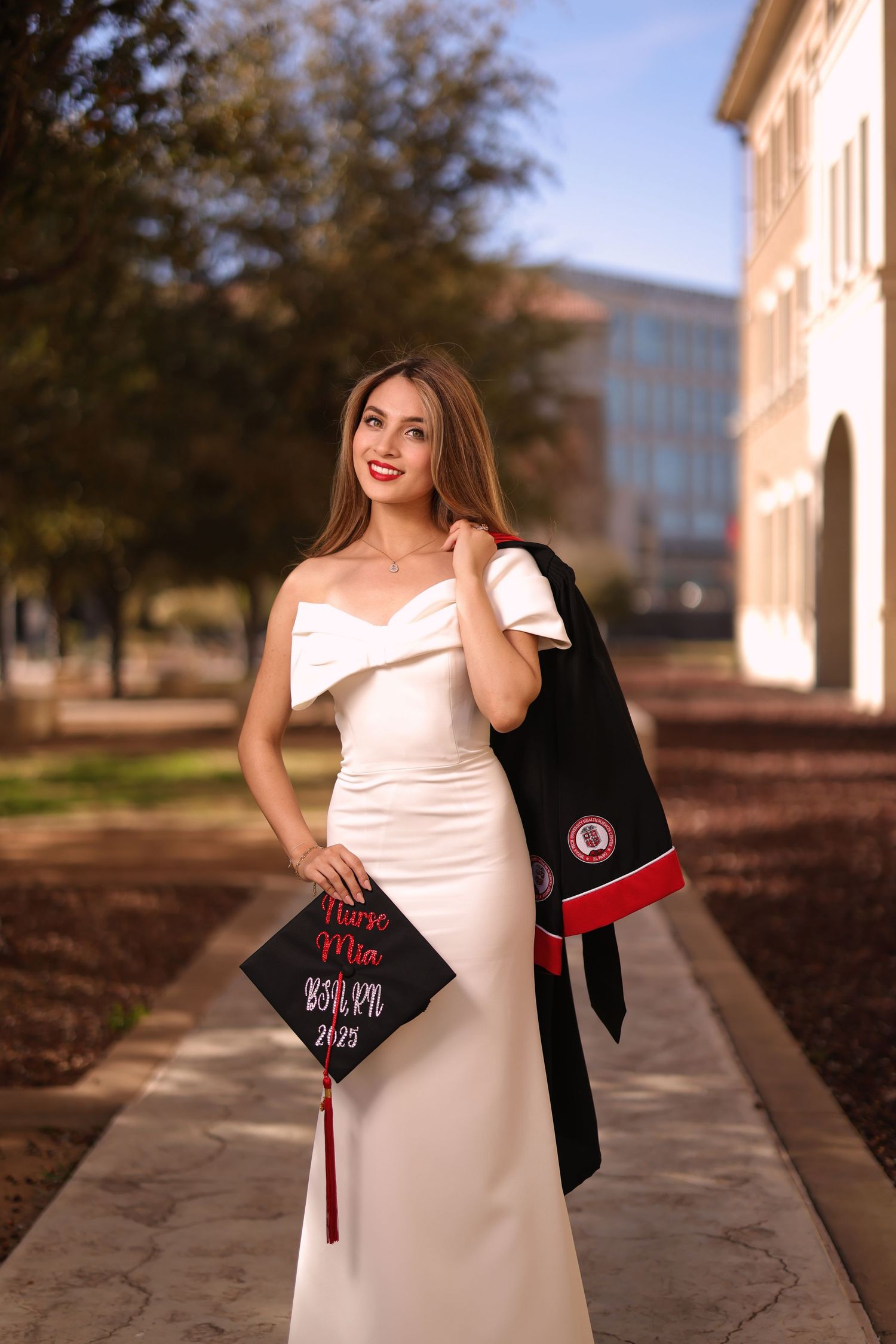 A graduate in a white formal dress holds a decorated graduation cap and black gown on a university campus walkway.