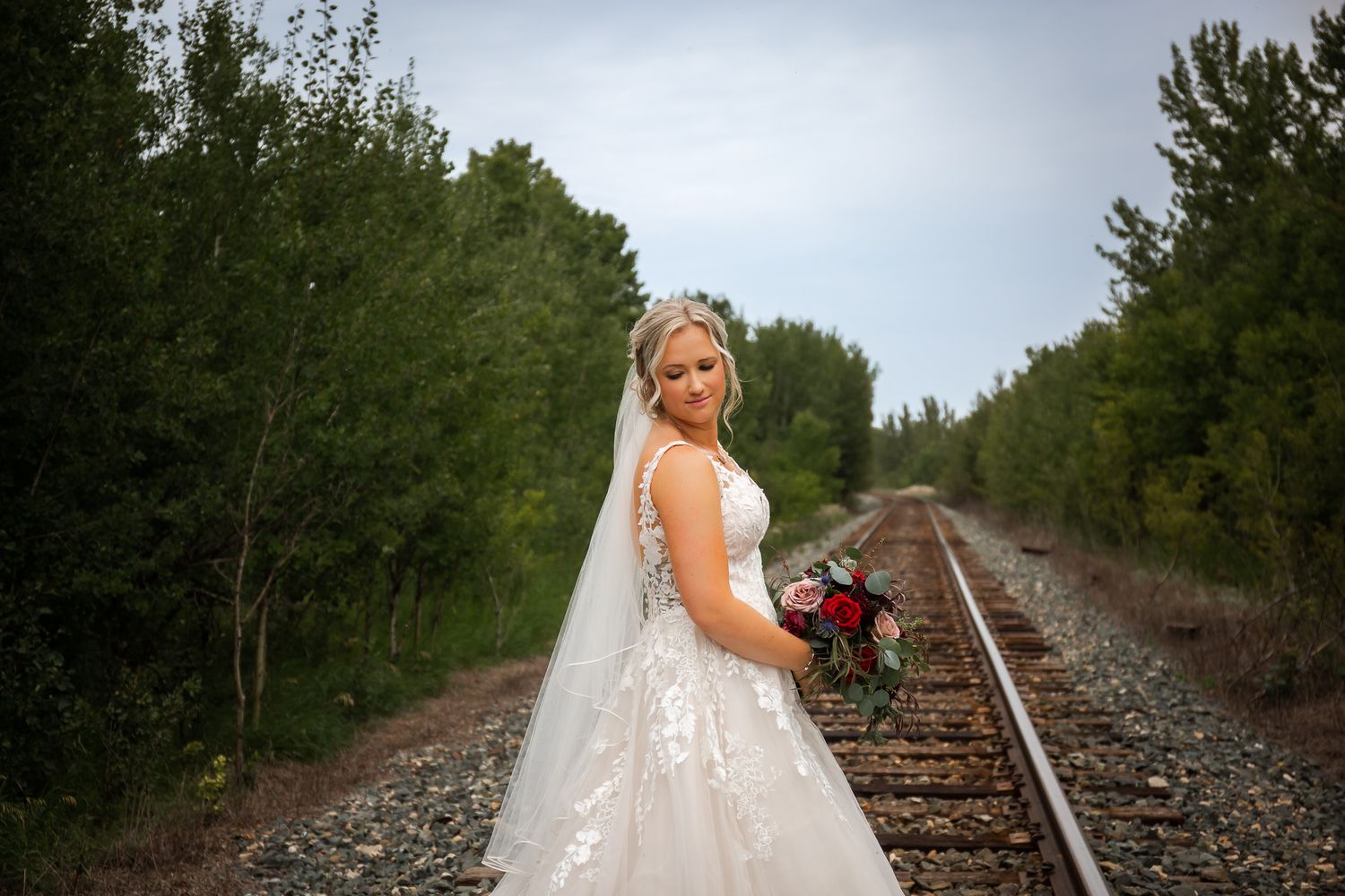 A bride in white dress poses with bouquet on railroad tracks surrounded by forest.