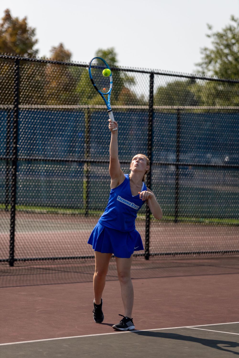 Tennis player in blue uniform serves the ball on outdoor court during sunny day.