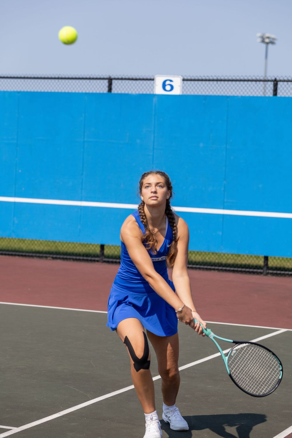 Tennis player in blue uniform prepares to serve with yellow ball tossed high on outdoor court.