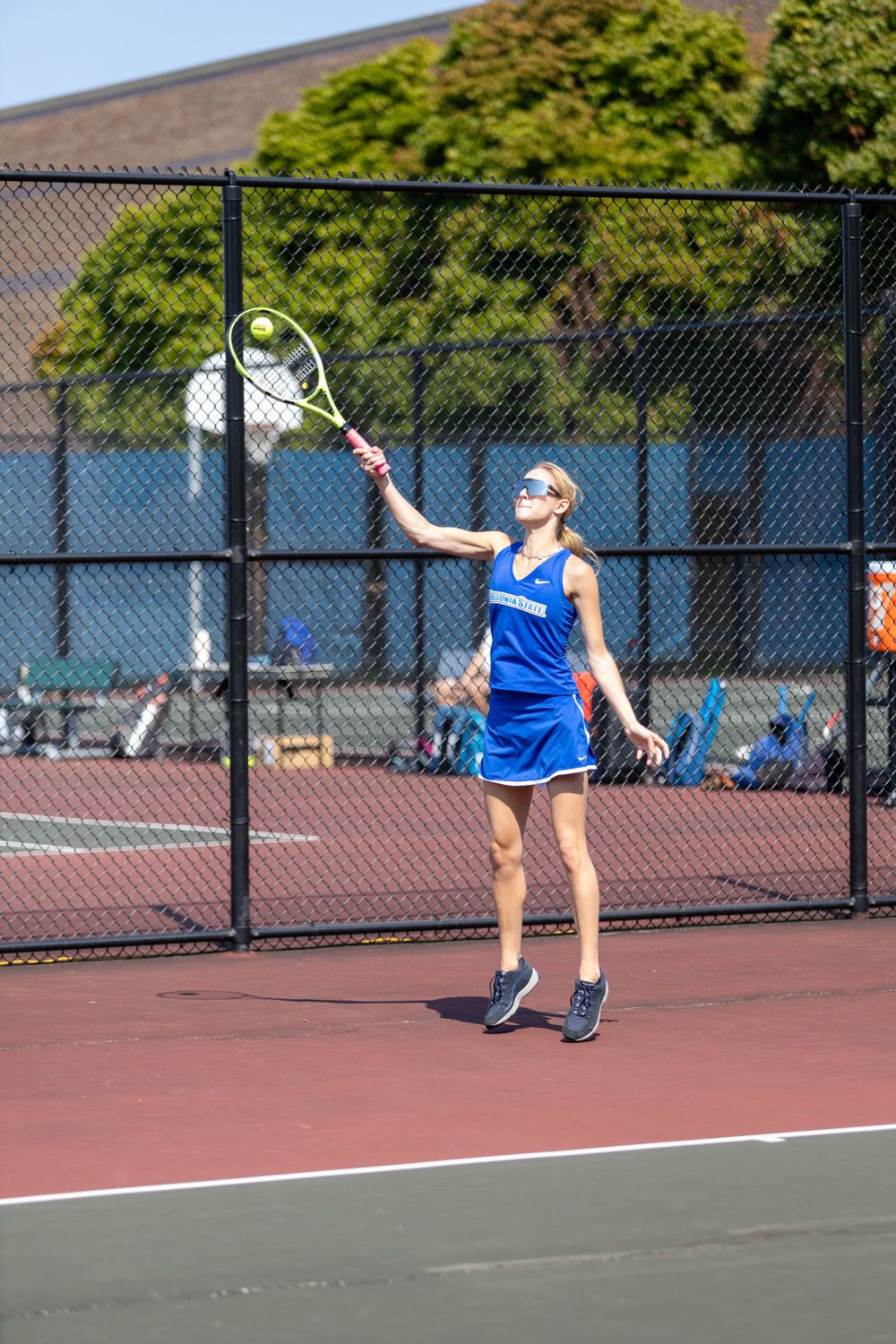 Tennis player in blue dress uniform serves ball on outdoor court with chain link fence background.