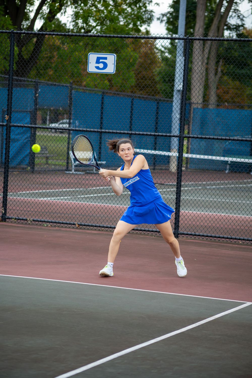 Tennis player in blue uniform stretches low to return a shot on outdoor court.