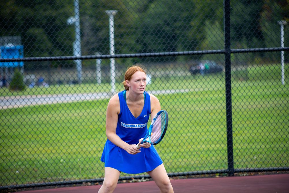 Player in royal blue uniform crouches low to make a tennis return shot.