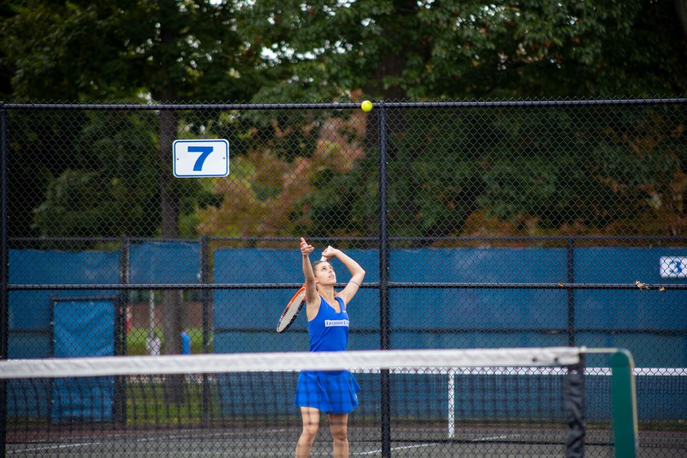 Tennis players compete on outdoor courts with spectators watching from behind fence.