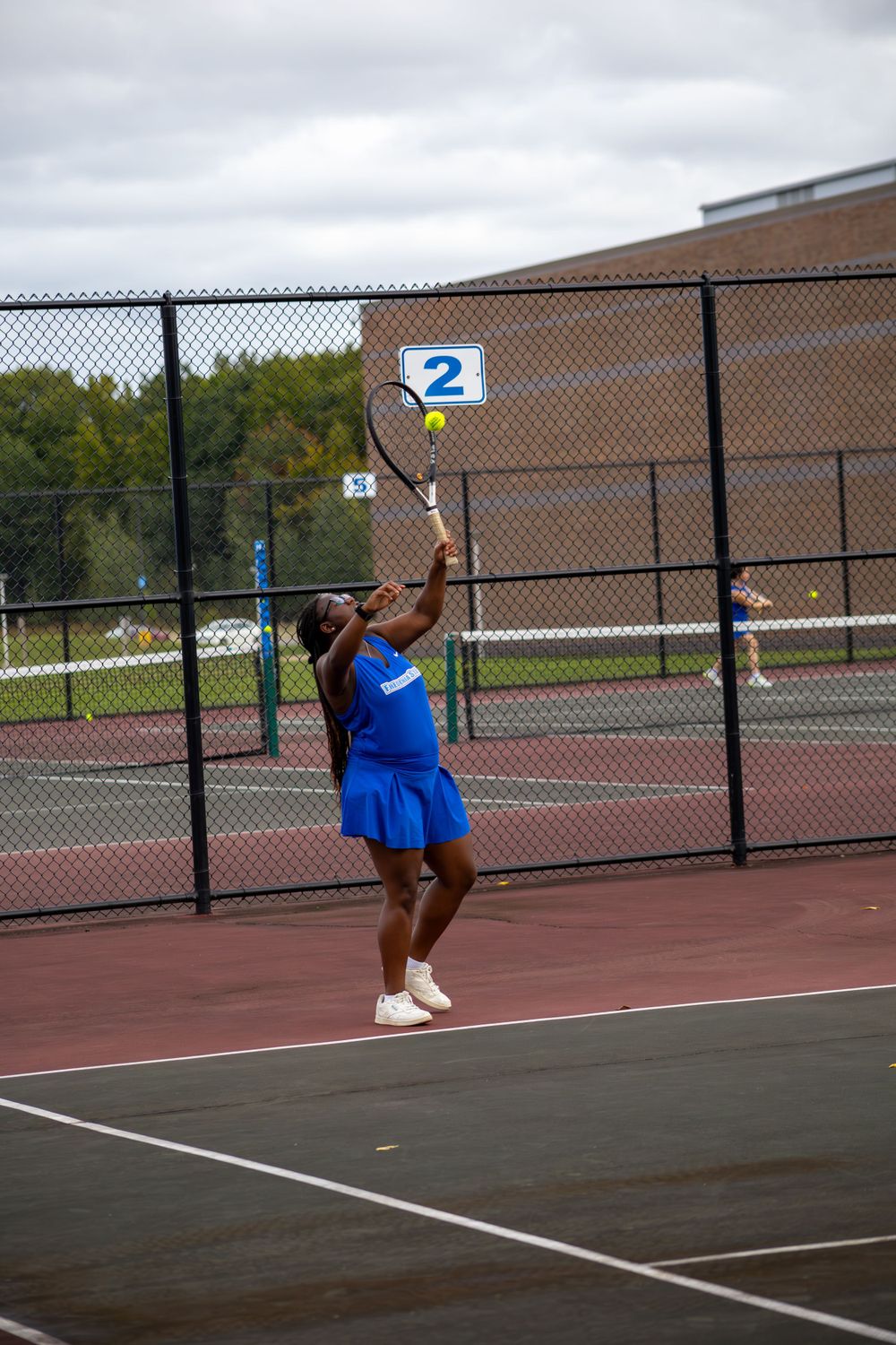 Player in bright blue uniform serves tennis ball on outdoor court.