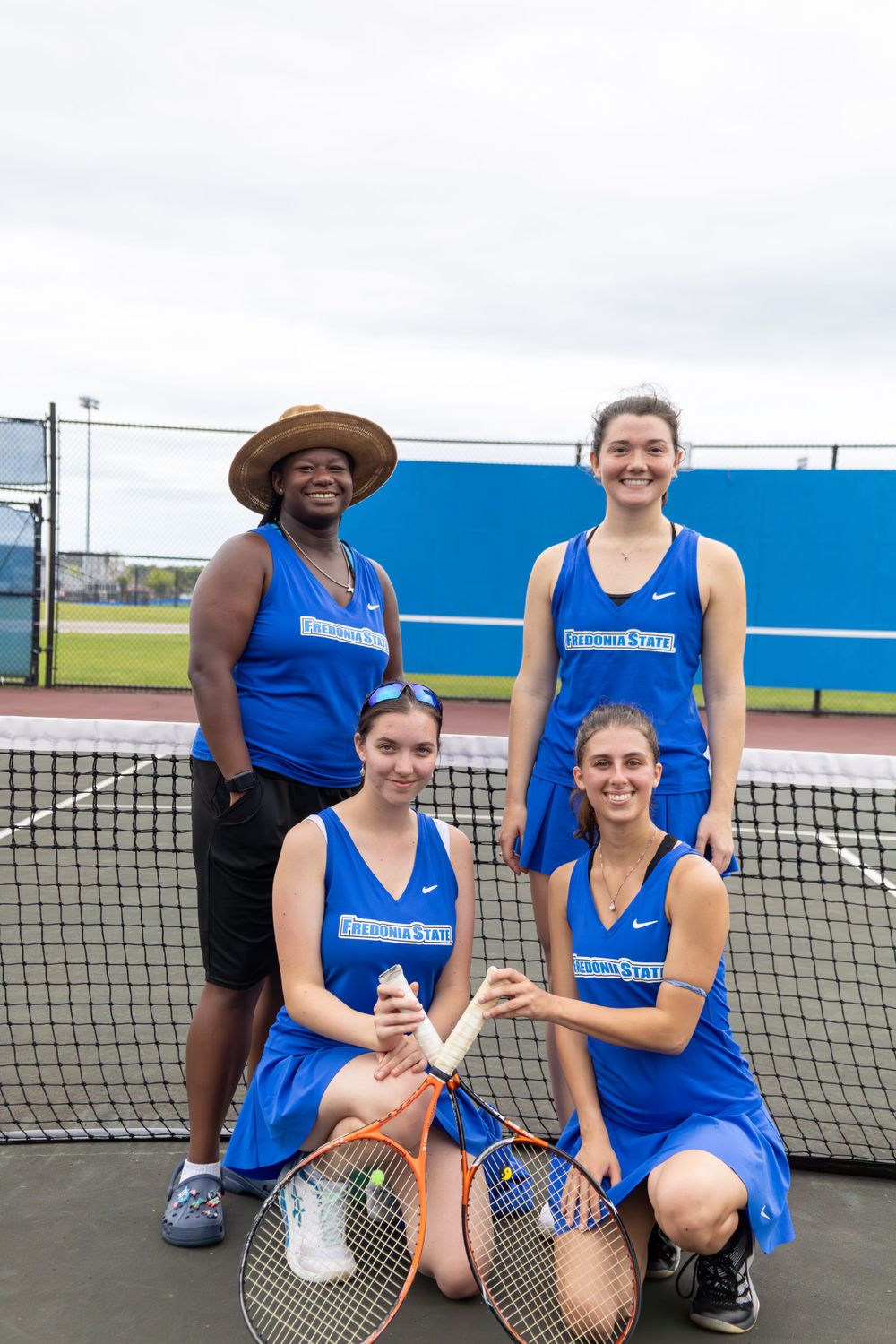 Tennis team members in blue uniforms pose together on outdoor courts with rackets.