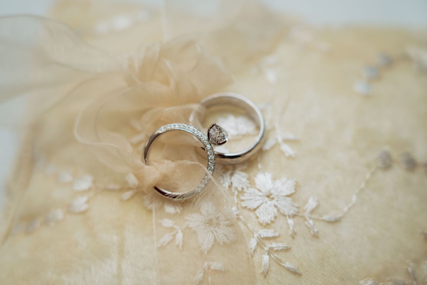 Two elegant silver wedding rings with diamond accents rest on a delicate cream colored lace background.
