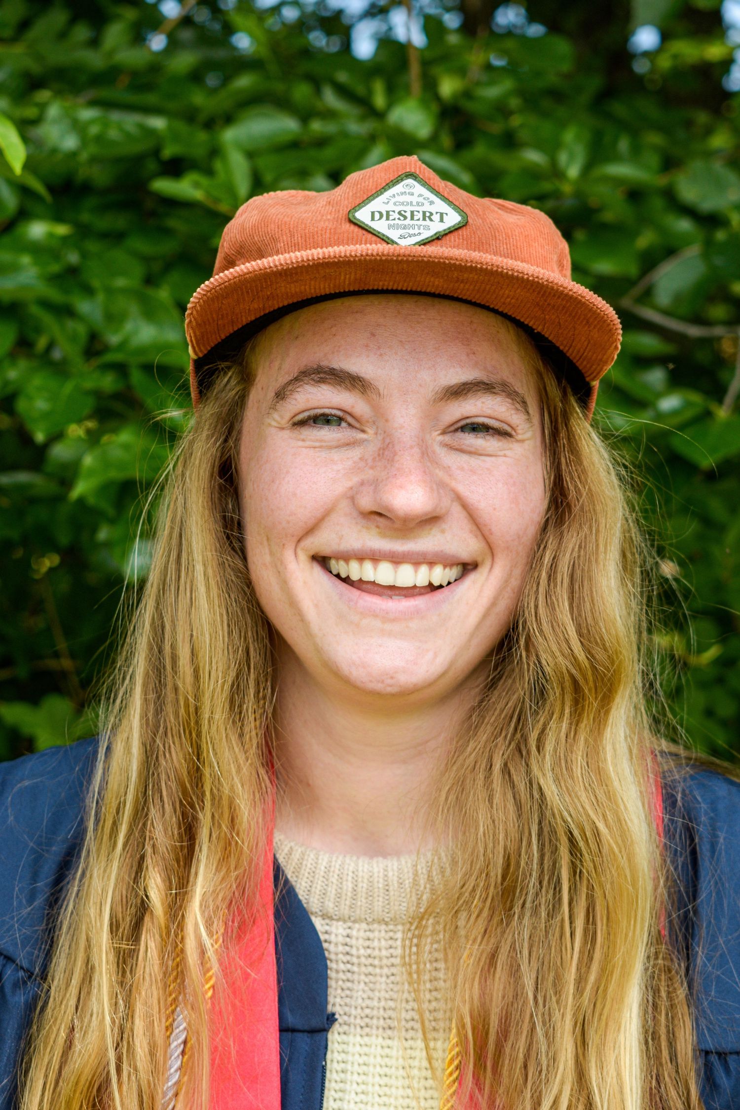 Person wearing an orange baseball cap smiles outdoors with green foliage in background.