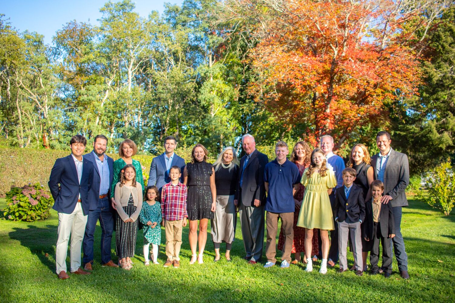 A large group poses together outdoors against a backdrop of autumn trees with vibrant orange and green foliage.
