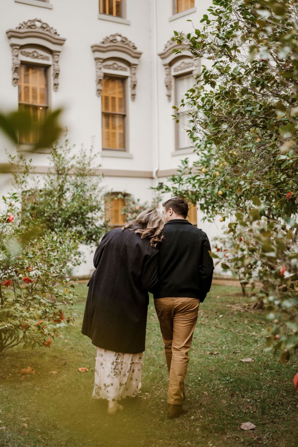 Couple embracing in a garden setting with ornate architecture in the background.