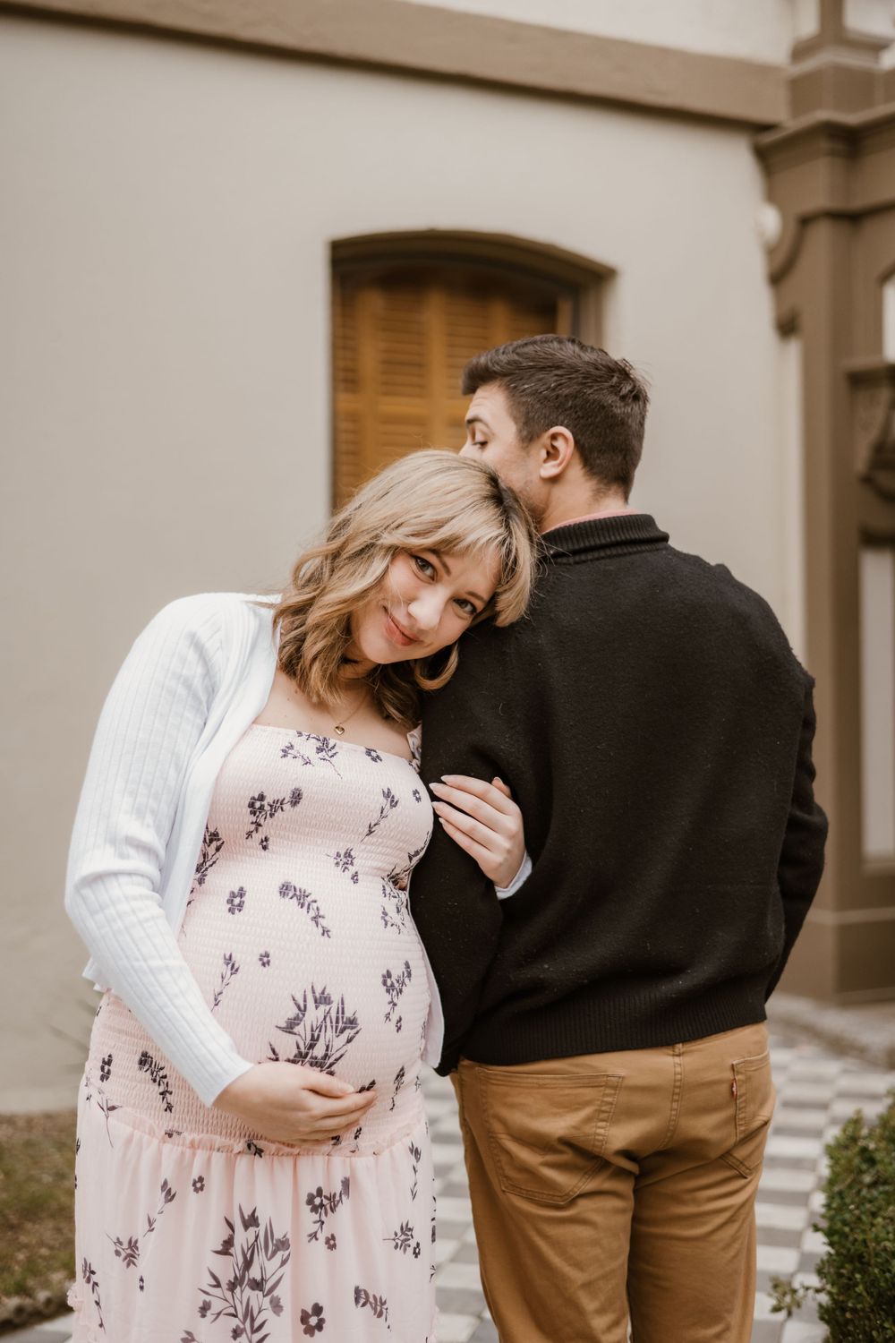 A tender maternity moment captured against a beige building with wooden shutters.