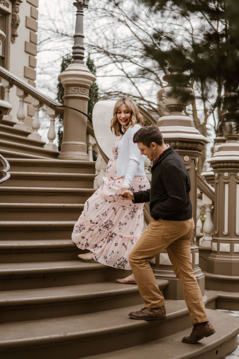 A couple ascends ornate stone steps while holding hands during a maternity photoshoot.