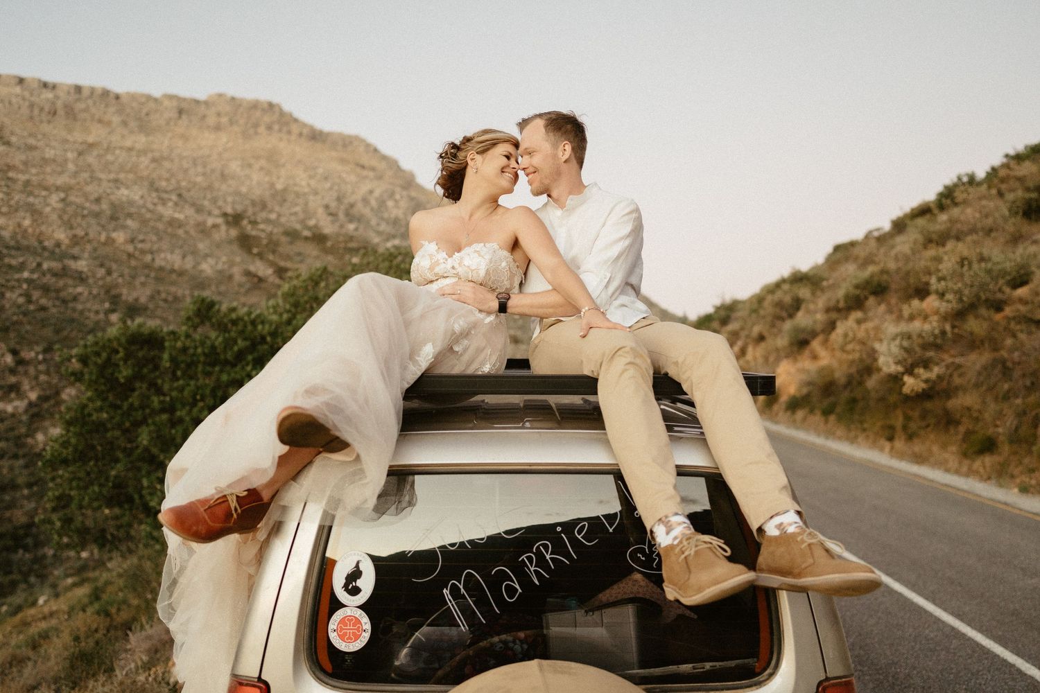 Couple in light wedding attire embraces on sandy dunes with mountain backdrop.