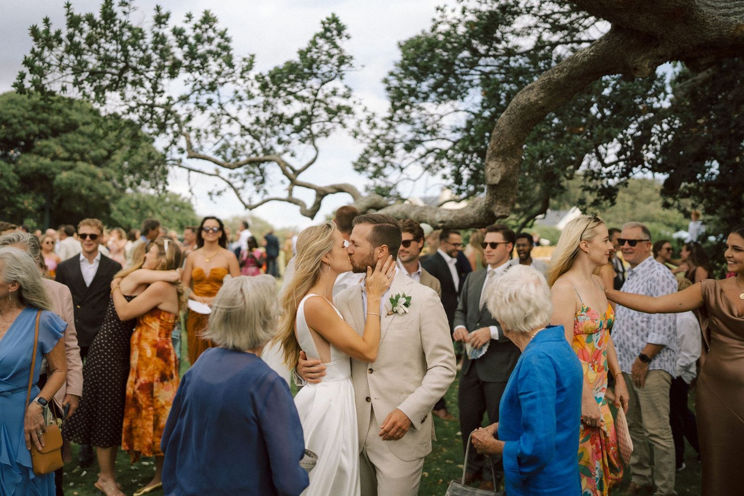 Couple in light wedding attire embraces on sandy dunes with mountain backdrop.